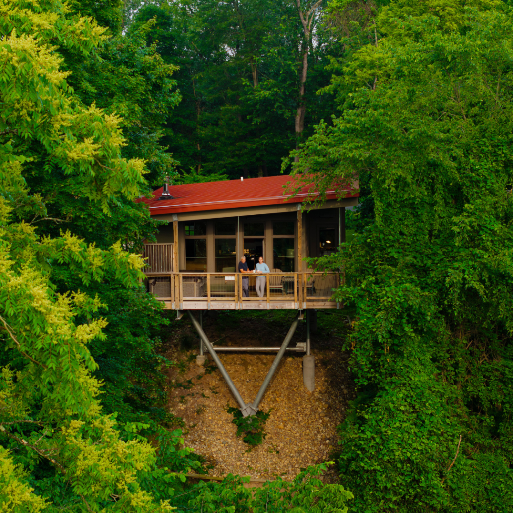 A wooden house on stilts surrounded by lush green trees, with two people sitting on the deck.