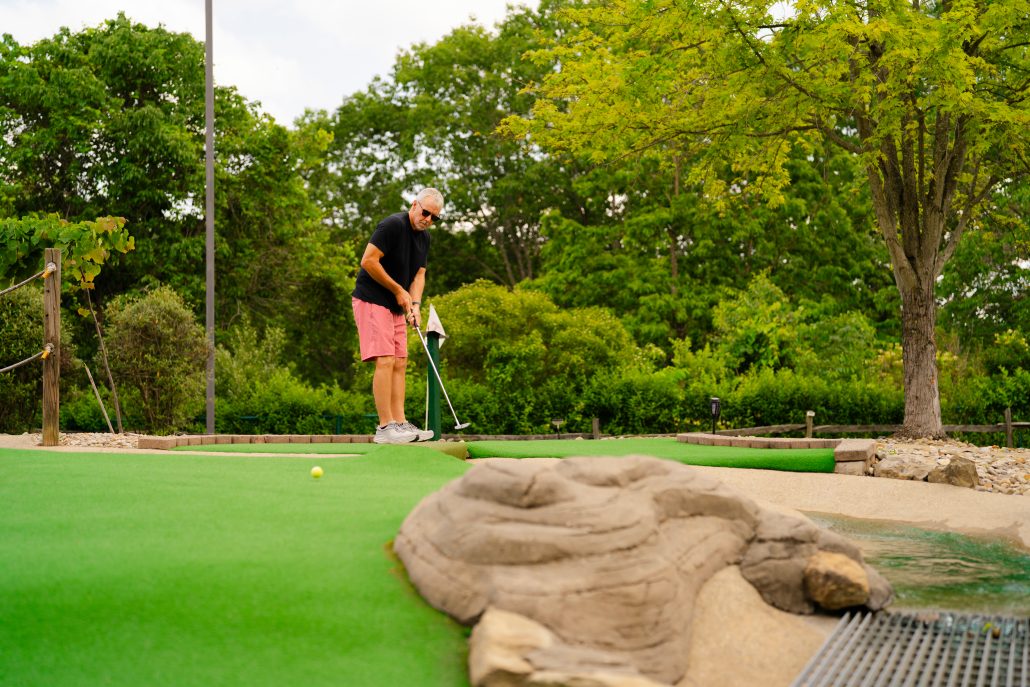 A man in a black shirt and pink shorts putts on a mini-golf course with green turf and trees in the background.