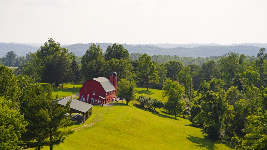 A red barn with a silo surrounded by green fields and trees under a bright sky.