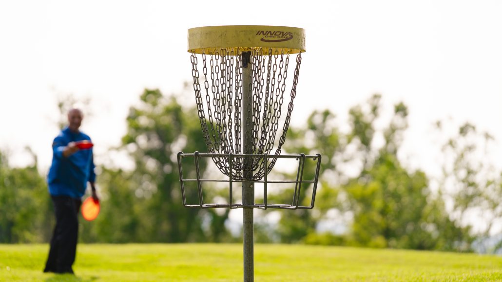 Disc golf basket with chains, labeled "INNOVA DISC GOLF," in a grassy area with a person throwing a disc in the background.