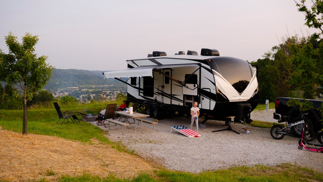 A camper trailer parked on gravel with a child playing, picnic table, and an American flag cornhole board.