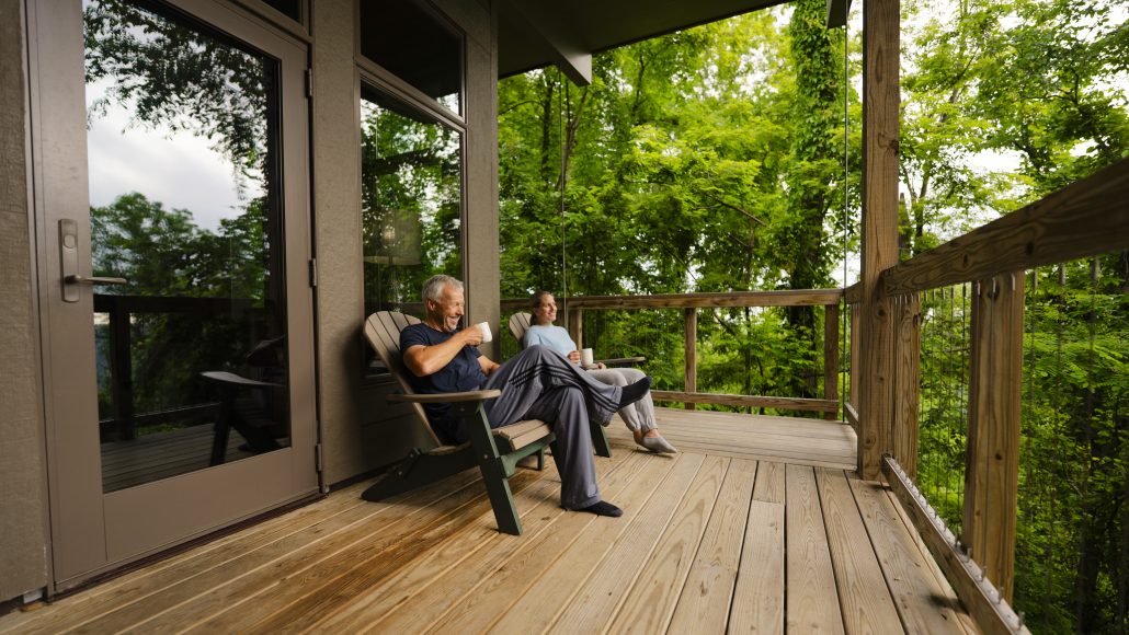 Two people sitting on a wooden deck surrounded by greenery, each holding a cup.