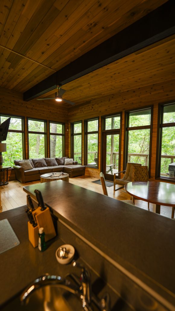 A cozy living area with large windows, a brown sectional sofa, and a round dining table, surrounded by greenery.