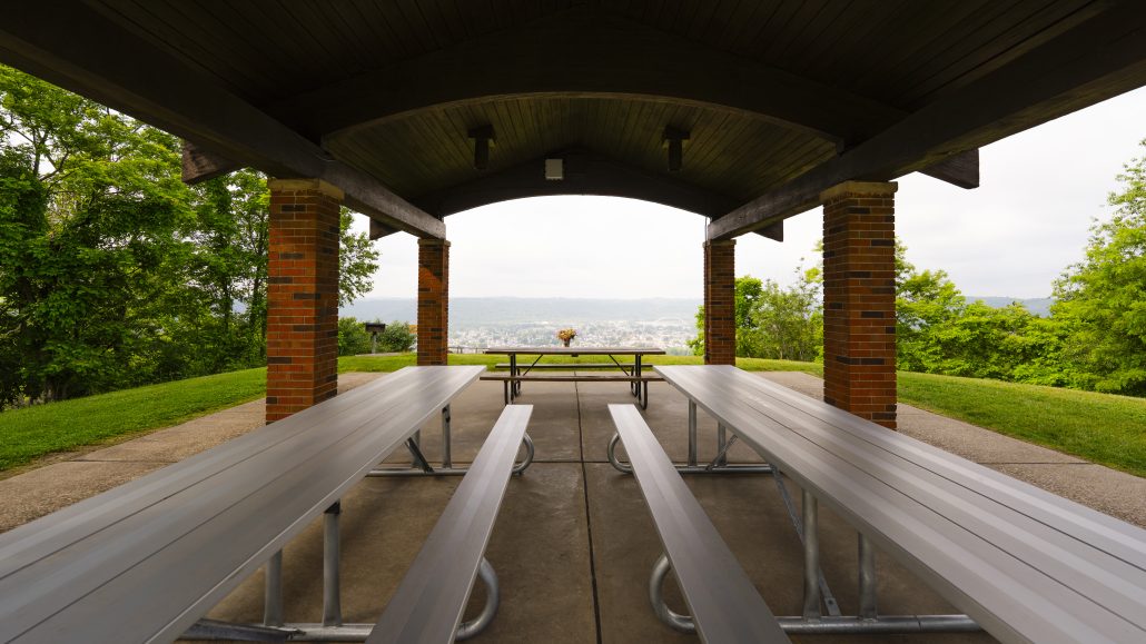 A covered picnic area with metal benches and a view of trees and hills in the background.