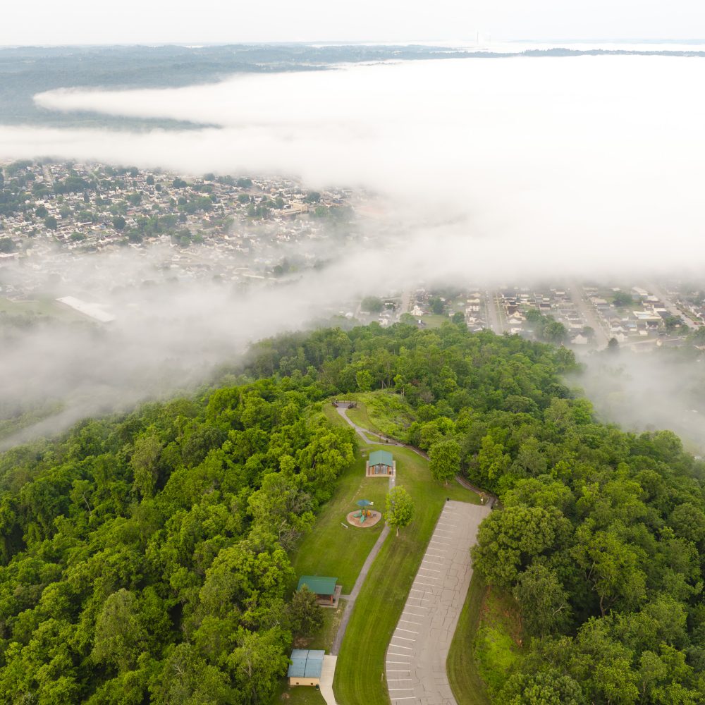 Aerial view of a green hillside with fog, overlooking a town and valley below.