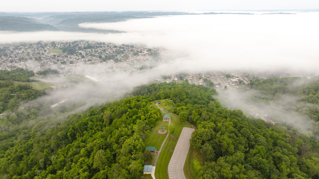 Aerial view of a green hillside with fog, overlooking a town and valley below.