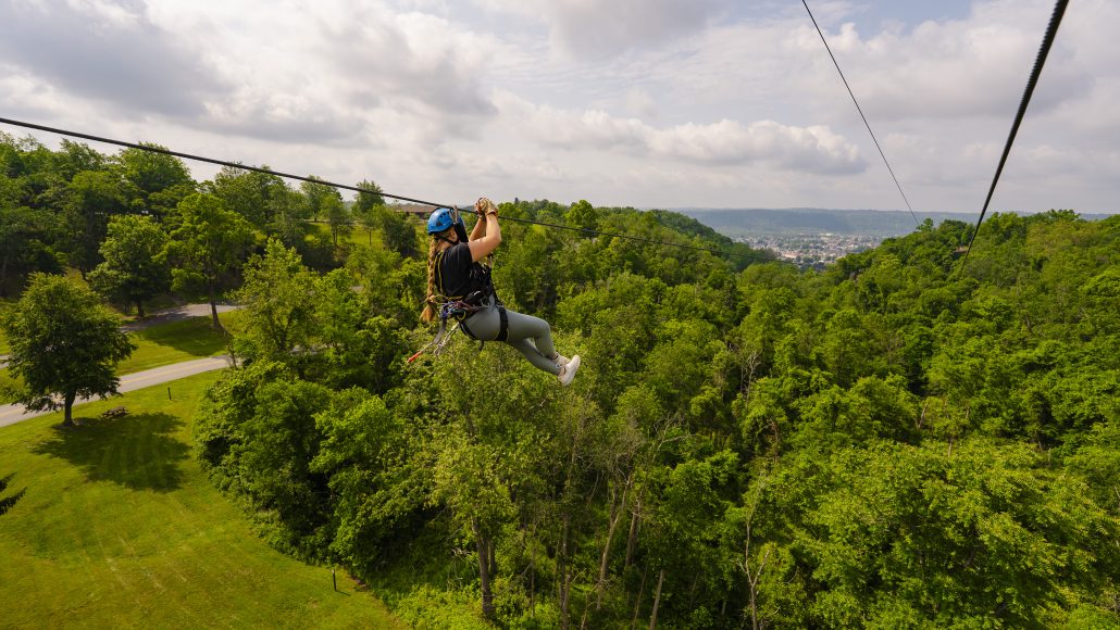 A person zip-lining through a green landscape with trees and a cloudy sky in the background.