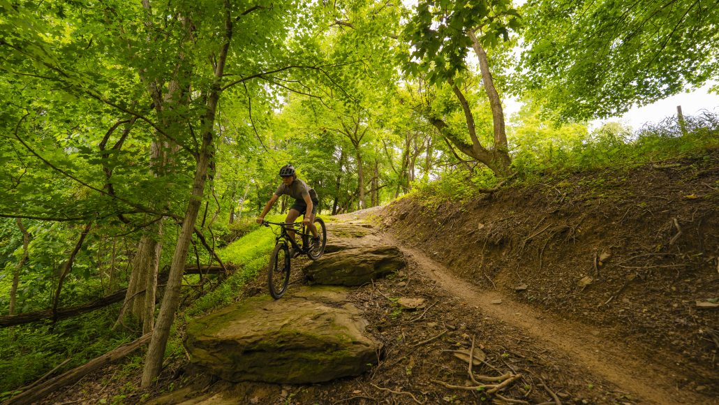 A person riding a mountain bike on a rocky trail surrounded by green trees.