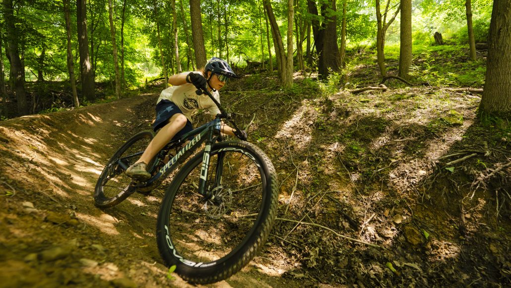 A person riding a mountain bike on a dirt trail surrounded by green trees. The bike has a "Specialized" logo.