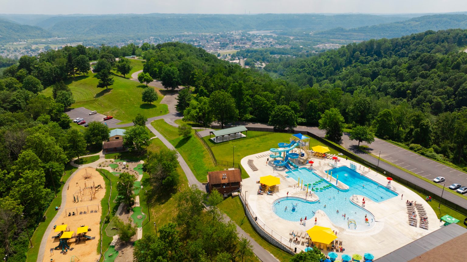 Aerial view of a swimming pool area with water slides, surrounded by green hills and a parking lot.