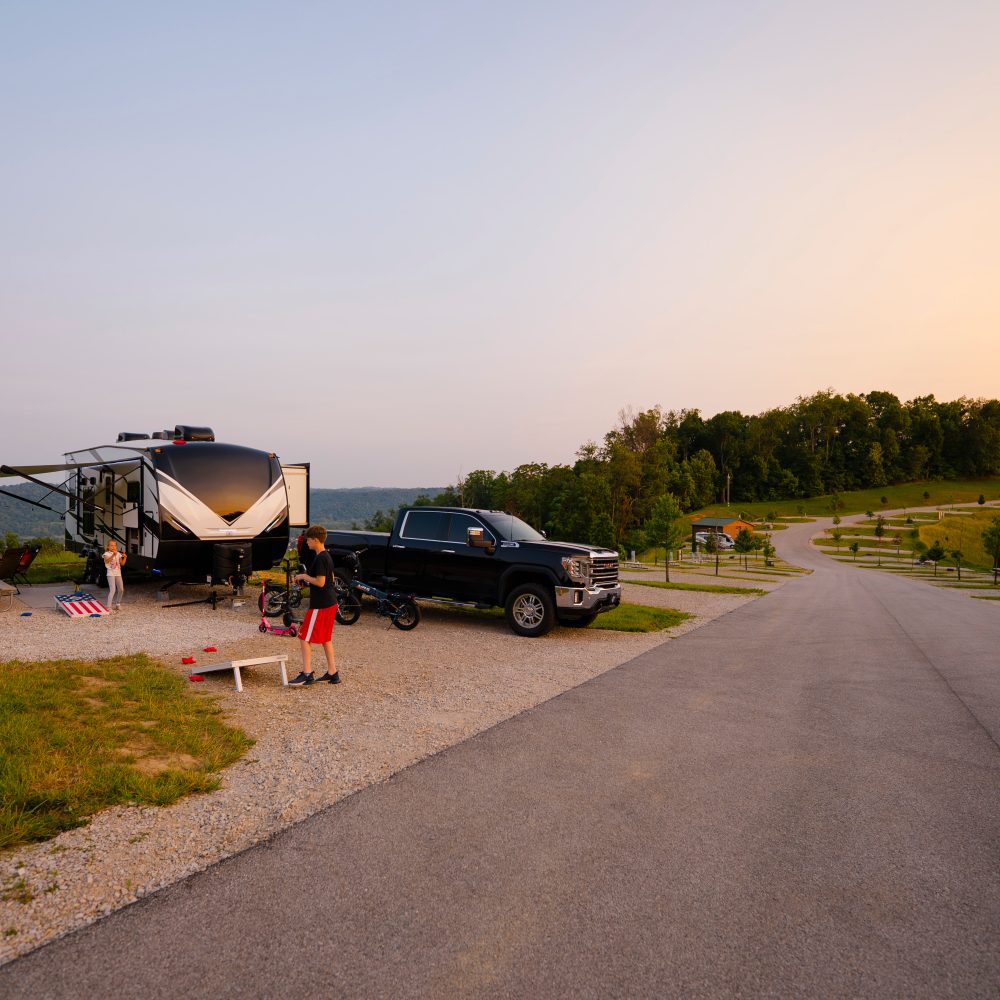 A camper and a black truck parked on gravel, with a child playing nearby and a scenic road in the background.