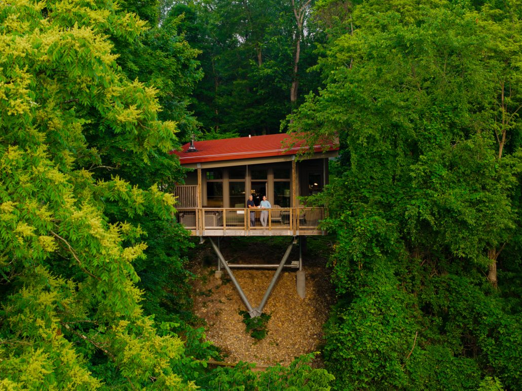 A modern house on stilts surrounded by lush green trees, with two people on the deck.