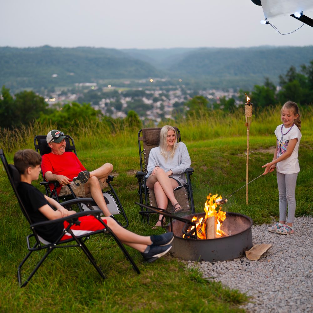 A family sits around a campfire, with a girl roasting marshmallows and a scenic view in the background.