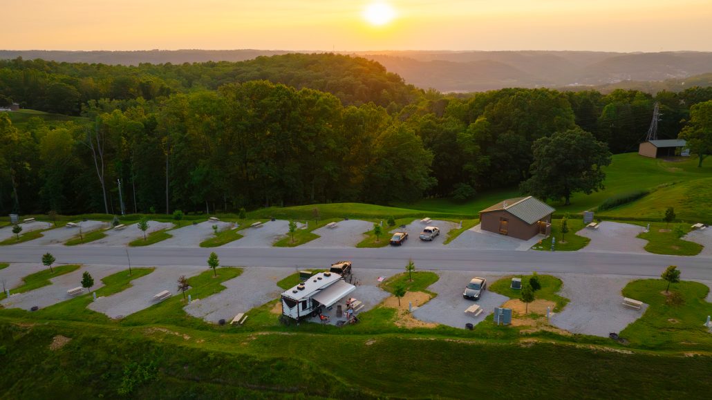 Aerial view of a campground with RVs, cars, picnic tables, and a restroom building at sunset.