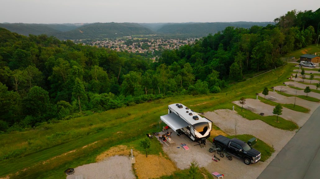 A camper and black truck parked on gravel near a green hillside, overlooking a valley and small town.