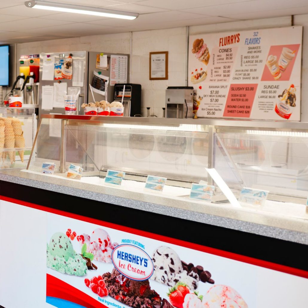 Ice cream shop counter with Hershey's logo, menu board, and ice cream flavors displayed in glass cases.