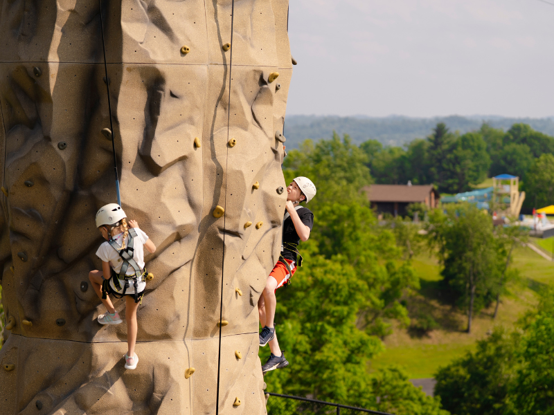 Two children climbing a rock wall, one in a white helmet and harness, the other in a black shirt and shorts.